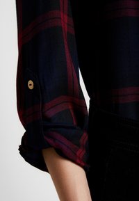 Een geruite shirtmouw in marineblauw, rood en zwart met een herringbone textuur. Het heeft een opgerolde manchette en een gouden knoopdetail op het lusje.