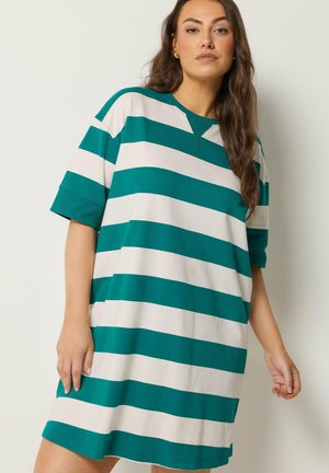 Kvinde med langt brunt hår iført en teal- og hvid bred horisontalstribet oversized t-shirt-kjole, stående foran en ensartet lys væg.