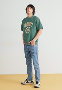 Sweatshirt verde com design gráfico, jeans azuis e ténis multicoloridos. Tecido suave, corte descontraído e design casual.