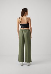 Fekete műbőr crop top, vékony pántokkal, laza, olajzöld, bő szárú nadrággal párosítva, ami rugalmas derékkal rendelkezik. Fehér sportcipők látszódnak.