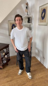 Νέος άντρας με ριγέ λευκό πουκάμισο και μαύρο παντελόνι στέκεται σε ένα δωμάτιο με ξύλινο πάτωμα, μοντέρνα τέχνη, ραφιέρες και ξύλινο μπαρ-καμπινέτο.