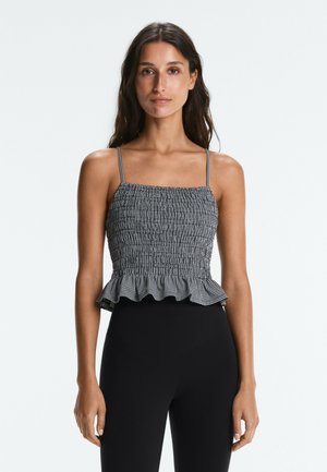 Žena má na sebe čierno-biele kockované crop top so sťahovaním, tenkými ramienkami a nariaseným lemom, v kombinácii s čiernymi nohavicami s vysokým pásom.