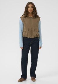 Kvinde med krøllet hår iført en brun ærmeløs vest over en lyseblå trøje, mørke jeans og brune sko, der står mod en hvid baggrund.