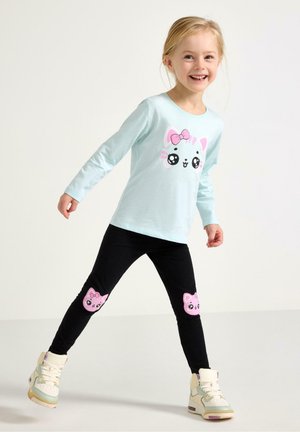 Smilende ung pige iført lyseblå langærmet skjorte og sorte leggings med lyserøde katteansigter på knæene, stående med benene spredt.