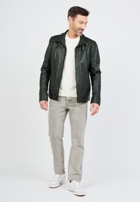 Groene leren jas met ritsen, ribbeltextuur en aangeknoopte kraag; gedragen over een witte trui, gecombineerd met grijze jeans en witte sneakers.