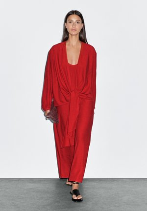 Femme portant un haut rouge à manches longues noué à la taille, une jupe longue assortie, tenant une pochette texturée et des sandales noires, marchant sur un sol gris.