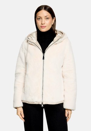 Frau trägt eine cremefarbene, flauschige Jacke mit Reißverschluss und Kapuze über einem schwarzen Rollkragenpullover und schwarzen Hosen, steht vor einem einfarbigen Hintergrund.