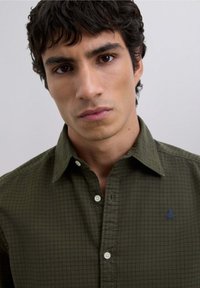 Dark green checkered shirt with a pointed collar, featuring white buttons and a small blue logo embroidered on the chest.