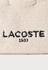 LACOSTE logo u crnoj veznoj špici na mekoj, bež, teksturiranoj pozadini, s prikazom godine "1933" ispod naziva brenda.