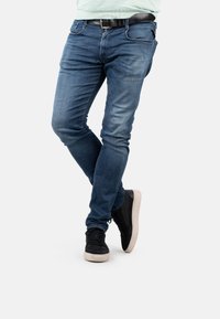 Jeans de mezclilla azul con un corte ajustado, que presentan un ligero desgaste y bolsillos frontales, combinados con zapatillas negras con una suela blanca.