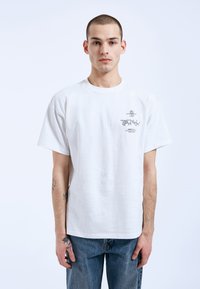 Bílý bavlněný tričko s volným střihem, krátkými rukávy a potiskem na levé části hrudníku. Nosené s modrými džíny.