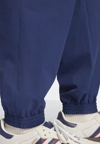 Námořnické joggingové kalhoty s elastickými manžetami, vyrobené z hladkého materiálu. V kombinaci s bílými teniskami s modrými a červenými akcenty.