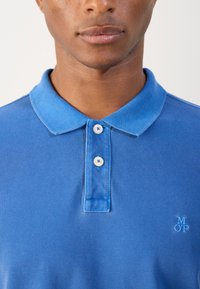 Blue polo shirt with a textured fabric, two white buttons, and a small embroidered logo on the chest. Classic collar design.