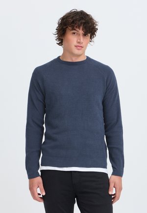 Jeune homme aux cheveux bouclés portant un pull bleu par-dessus une chemise blanche et un pantalon noir, debout devant un fond clair uni.