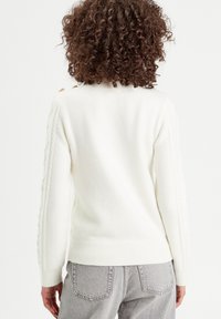 Pull en tricot écru avec des poignets et un ourlet côtelés, orné d'un motif en jersey le long des manches et de boutons décoratifs à l'épaule.