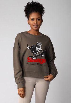 Femme portant un sweat-shirt marron avec un graphique d'un chat sautant par-dessus un skateboard et le mot "Savage" en texte rouge, assorti de pantalons beiges.