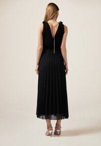 Zwarte geplooide maxi-jurk met textuur in het lijfje, V-rug en gouden kettingdetail. Gekocht met zilveren gesp sandalen, die een sheer zoom laten zien.