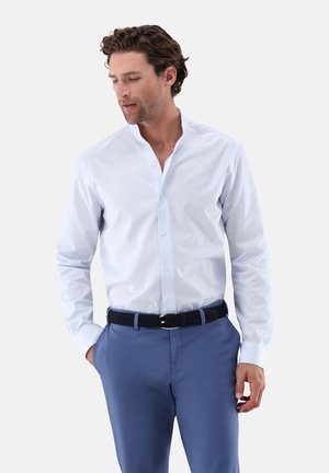 Mann mit lockigem Haar, der ein hellblaues Hemd mit Knöpfen und blaue Hosen mit einem dunklen geflochtenen Gürtel trägt, Hände in den Taschen, blickt nach unten.