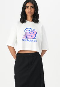 Balta apgriezta t-kreklu ar pārspīlētām īsām piedurknēm, uz kura ir krāsains "New Balance" logo zila un rozā krāsā, kopā ar melnu svārku ar elastīgu jostasvietu.