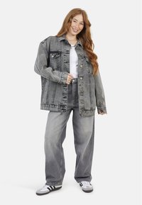 Veste en denim grise surdimensionnée avec fermeture à bouton et poches, associée à un jean large gris clair et des chaussures de sport noir et blanc.