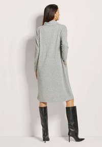 Graues Strickkleid mit langen Ärmeln und Rollkragen, ausgestattet mit seitlichen Taschen. Kombiniert mit knielangen schwarzen Leder-Stilettos.