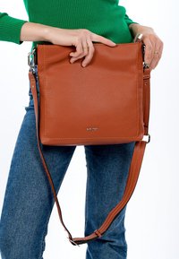 Brūna ādas plecu soma ar strukturētu virsmu, regulējamu siksnu un sudraba detaļām. Soma ir veidota minimālistiskā stilā.