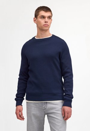 Giovane uomo che indossa un maglione a coste blu navy sopra una camicia bianca e jeans grigio chiaro, in piedi davanti a uno sfondo bianco semplice.
