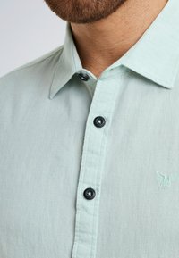 Hellgrünes Leinenhemd mit Button-Down-Kragen und schwarzen Knöpfen. Dezentes, auf der Brust gesticktes Adleremblem. Sanfte Textur.