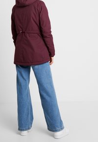 Eine Person trägt eine bordeauxroteJacke, weite blaue Jeans und weiße Sneakers und steht vor einem schlichten hellgrauen Hintergrund.