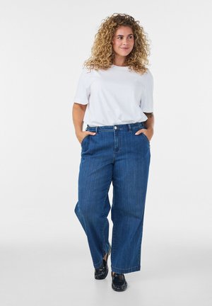 Kvinde med krøllet hår iført hvid T-shirt og blå vide jeans med hænderne i lommerne, stående på en ensfarvet hvid baggrund.