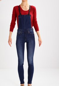 Femme portant une salopette en denim bleu foncé avec des bretelles noires et un haut rouge à manches longues, debout devant un fond blanc.