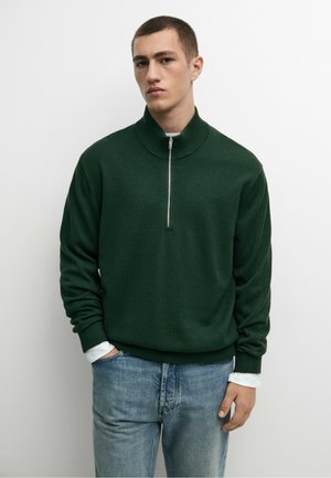 Junger Mann mit kurzen Haaren, der einen dunkelgrünen Reißverschluss-Pullover über einem weißen Hemd und hellblauen Jeans trägt, vor einem einfarbig weißen Hintergrund steht.