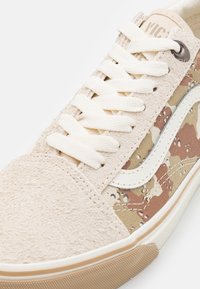Nahaufnahme eines beigen Wildleder-Sneakers mit weißen Schnürsenkeln, camouflagefarbenen Stoffseiten, weißem Wellenstreifen und Gummisohle in Kaugummifarben.
