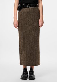 Jupe maxi en tricot marron avec des côtes verticales, associée à une ceinture noire avec une boucle circulaire et des poches. Portée avec des chaussures de sport noires.