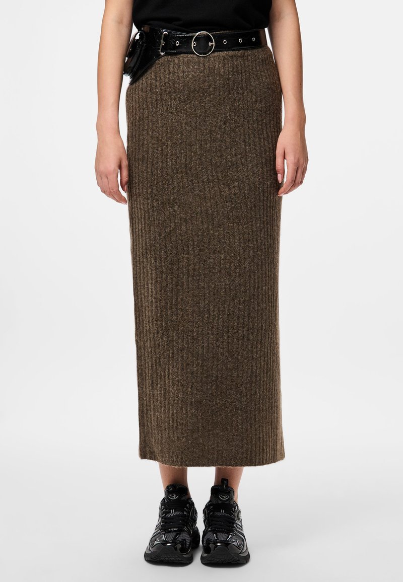 Jupe maxi en tricot marron avec des côtes verticales, associée à une ceinture noire avec une boucle circulaire et des poches. Portée avec des chaussures de sport noires.