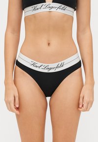 Zwarte stringonderbroek van een zachte stof, met een brede witte tailleband. De tailleband heeft "Karl Lagerfeld" in cursieve letters.