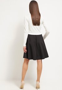 Vrouw met lang steil haar, gekleed in een witte longsleeve top, een zwart knielang rokje en beige hoge hakken, staand met de rug naar de kijker gekeerd.