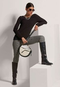 Schwarzes, geripptes Langarmoberteil, gemusterte schwarz-weiße Hose, kniehohe schwarze Lederstiefel, runde Handtasche in Creme und Schwarz, Sonnenbrille.
