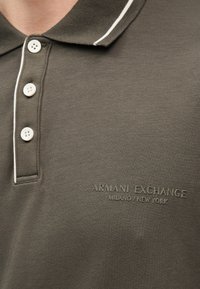 Χακί πράσινη μπλούζα πόλο με λευκό περίγραμμα στο γιακά και τα κουμπιά, με ανάγλυφο λογότυπο "Armani Exchange Milano / New York" στο στήθος.
