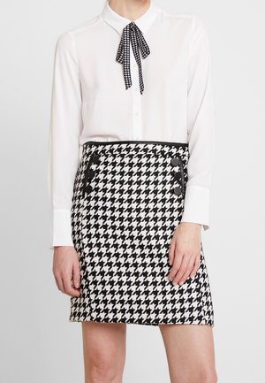 Une femme porte un chemisier blanc à manches longues avec un nœud papillon noir à motif pied-de-poule et une jupe assortie pied-de-poule noire et blanche avec des boutons noirs.