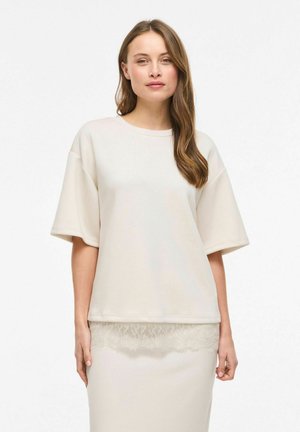 Kobieta o długich brązowych włosach, ubrana w luźną kremową bluzkę z krótkim rękawem i dopasowaną spódnicę z koronkowym wykończeniem, stojąca na białym tle.