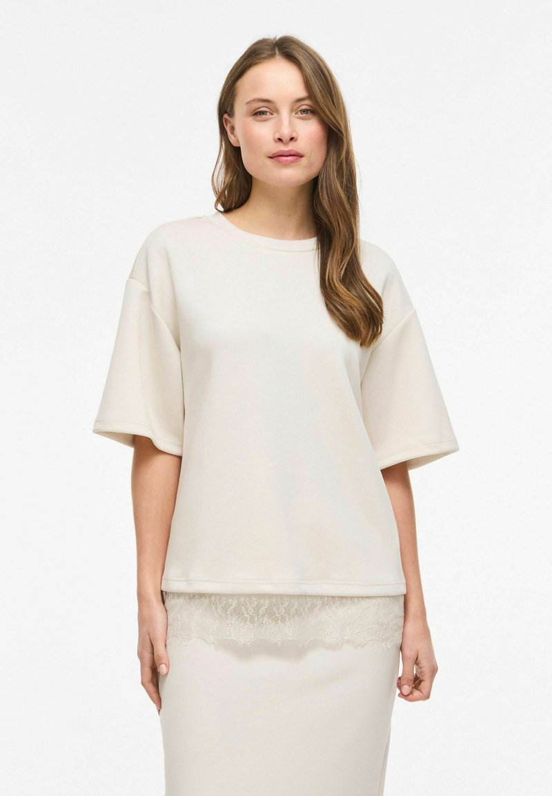 Femme aux longs cheveux bruns portant un haut ample à manches courtes crème et une jupe assortie avec bordure en dentelle, debout devant un fond blanc.