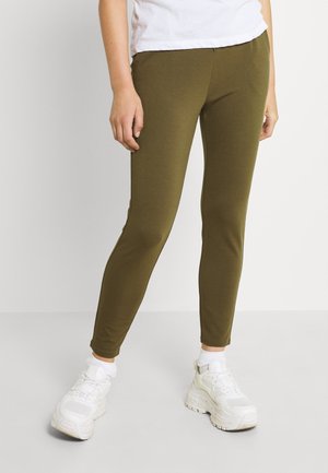 Leggings verde-oliva feitos de tecido elástico, apresentando um corte justo e sem costuras visíveis. Usados com ténis brancos e meias até ao tornozelo.
