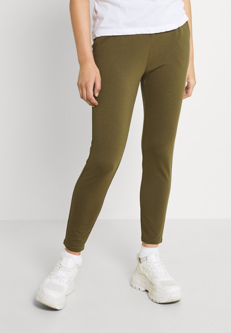 Leggings verde-oliva feitos de tecido elástico, apresentando um corte justo e sem costuras visíveis. Usados com ténis brancos e meias até ao tornozelo.