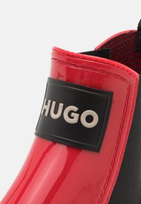 Botte de pluie en caoutchouc rouge avec un patch logo noir portant l'inscription "HUGO". Texture lisse avec une finition brillante et des accents de couleur contrastés.