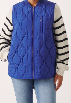 Osoba ubrana w niebieską pikowaną kamizelkę z przednim suwakiem na białym swetrze w czarne paski i jasnoniebieskie dżinsy, stojąca z jedną ręką w kieszeni.