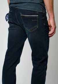 Tmavě modré džínové kalhoty s přiléhavým střihem, vybavené zipovou kapsou na zadní straně a jemnými šicími detaily. Texturovaná látka.