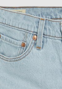 Jeans de denim azul claro con remaches de cobre, costuras en contraste y un bolsillo frontal. El material es suave, con un acabado ligeramente texturizado.