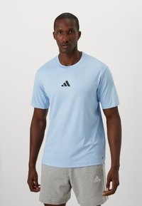 Lyseblå treningstrøye laget av teksturert stoff, med svart Adidas-logo sentrert på brystet. Korte ermer, rund halsdesign.