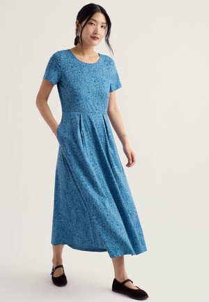 Femme debout avec les mains dans les poches, portant une robe bleue à manches courtes, longueur cheville, avec un discret motif floral et des ballerines noires.